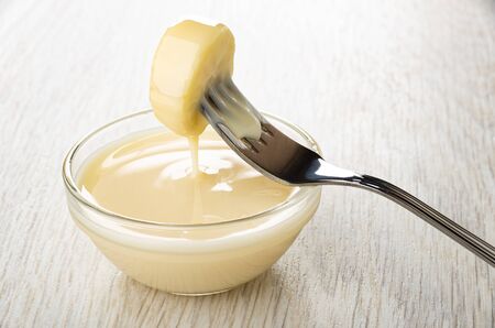 Piece of banana strung on fork on glass transparent bowl with condensed milk on wooden tableの写真素材