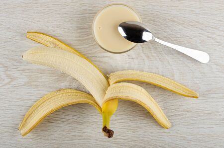 Peeled yellow banana, teaspoon on transparent glass bowl with condensed milk on wooden table. Top viewの写真素材