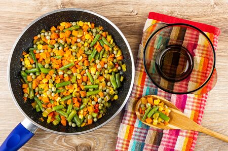 Fried vegetable mix in frying pan, vegetables on bamboo spoon, brown glass bowl on checkered napkin on wooden table. Top viewの写真素材