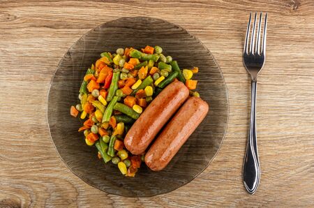 Grilled meat sausages with vegetable mix in black transparent plate, fork on wooden table. Top viewの写真素材