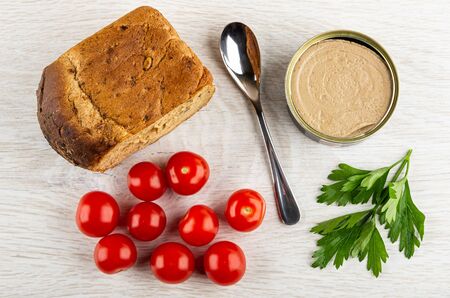 Loaf of bread, teaspoon, metallic jar with liver pate, red tomato cherry, leaves of parsley on wooden table. Top viewの写真素材