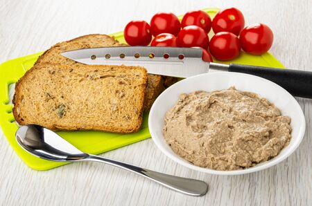 Pieces of bread, kitchen knife, tomato cherry on cutting board, teaspoon, white bowl with liver pate on wooden tableの写真素材