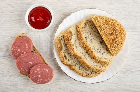 Sandwich with sausage, bowl with ketchup, slices of bread in white plate on wooden table. Top view