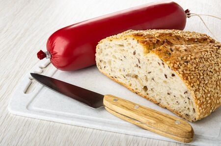 Sausage in polyethylene pack with rope, kitchen knife, piece of bread on plastic cutting board on wooden tableの写真素材