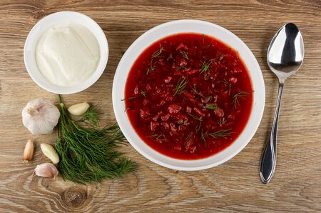Sour cream in bowl, dill, garlic, white plate with borscht, spoon on wooden table. Top view