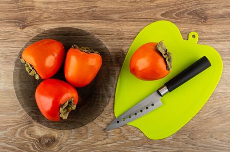 Fresh persimmons in black glass plate, permission and kitchen knife on green cutting board on wooden table. Top viewの写真素材