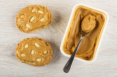 Two sandwiches with peanut butter, plastic container with peanut butter, teaspoon on light wooden...