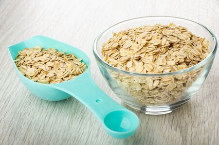 Blue plastic spoon with oat flakes, flakes in transparent glass bowl on wooden table