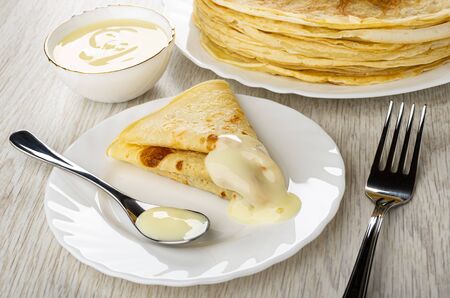 Bowl with condensed milk, stack of russian pancakes in dish, folded pancake, teaspoon with condensed...