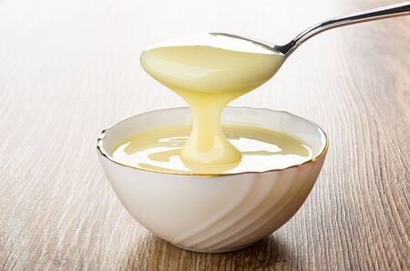 Pouring condensed milk from teaspoon in white glass bowl on wooden tableの写真素材