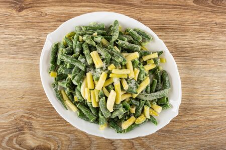 Frozen raw yellow and green beans in white glass dish on wooden table. Top view