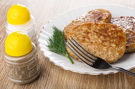 Salt and pepper shakers, fried pork cutlets and dill in white glass plate, fork on wooden table