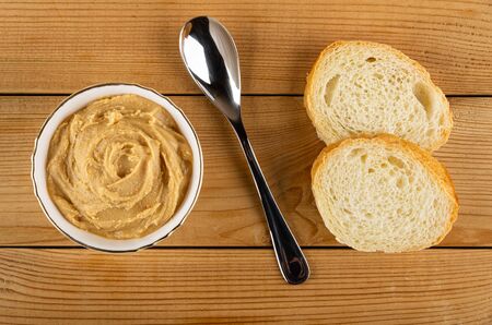 Peanut butter in white bowl, teaspoon, slices of wheat bread on wooden table. Top view