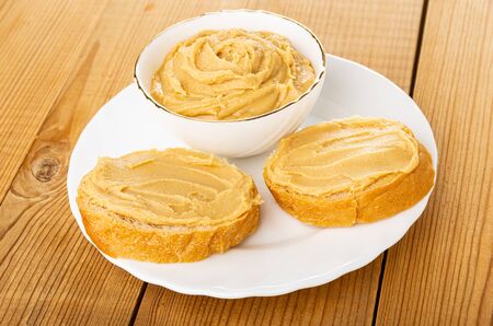 Peanut butter in white bowl, sandwiches in glass plate on wooden table