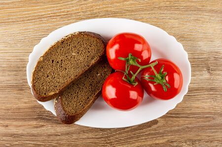 Slices of bread, fresh red tomatoes on branch in white dish on wooden table. Top view