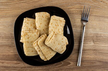 Few pancake rolls with filling in glass black plate, fork on wooden table. Top view