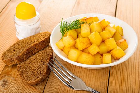 Salt shaker, slices of bread, white plate with cubes of fried potatoes, fork on wooden tableの写真素材