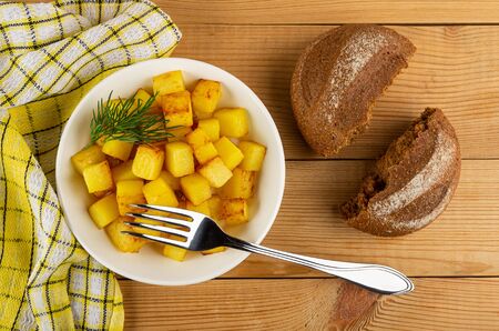 Checkered napkin, fork in white plate with cubes of fried potatoes, dill, pieces of bread on wooden table.の写真素材