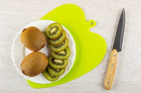 Slices of kiwi fruits, whole kiwi in white glass plate on cutting board, kitchen knife on light wooden table. Top viewの写真素材