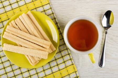 Heap of breadsticks with filling in saucer on checkered napkin, yellow cup with tea, teaspoon on...