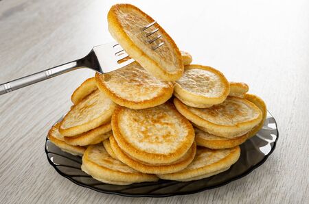 Pancake strung on fork above heap of homemade fried pancakes in gray transparent plate on wooden...