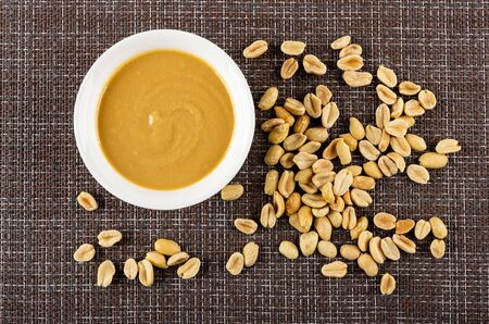 White glass bowl with peanut butter, scattered fried peanuts on brown mat. Top viewの写真素材
