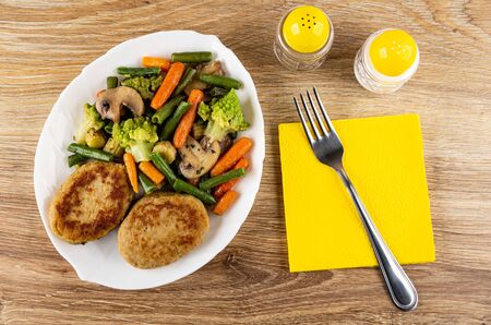 Fried spanish vegetable mix and turkey cutlets in white glass dish, fork on yellow napkin, salt and pepper shakers on wooden table. Top viewの写真素材