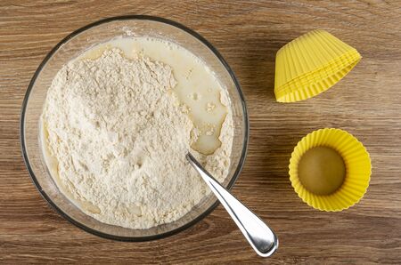 Transparent bowl with flour mix for muffins and milk, yellow silicone molds on wooden table. Top...