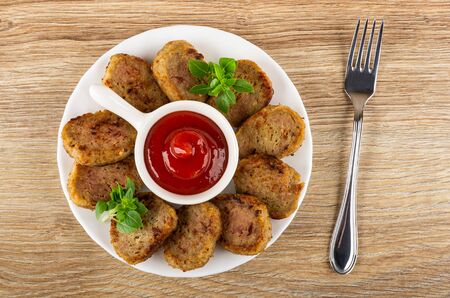 Tomato ketchup in sauceboat with handle, small chicken cutlets, basil in white glass plate, fork on...