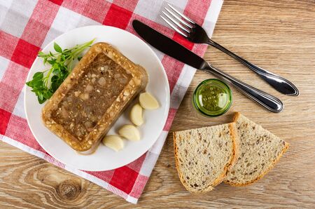Piece of meat jelly, basil, slices of garlic in white glass plate, kitchen knife and fork, pepper shaker on checkered napkin, slices of bread on wooden table. Top viewの写真素材