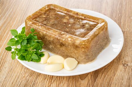 Piece of meat jelly, basil, slices of garlic in white glass plate on wooden table