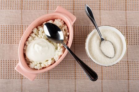Spoon in  pink glass bowl with defatted grained cottage cheese and sour cream, teaspoon in bowl with sugar on checkered napkin on table. Top viewの写真素材