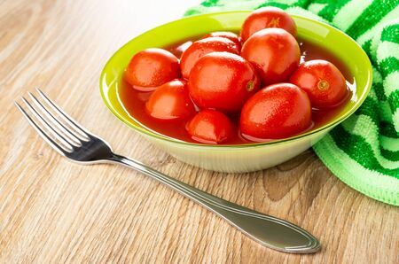Fork, green bowl with canned tomatoes in own juice, striped napkin on brown wooden tableの写真素材