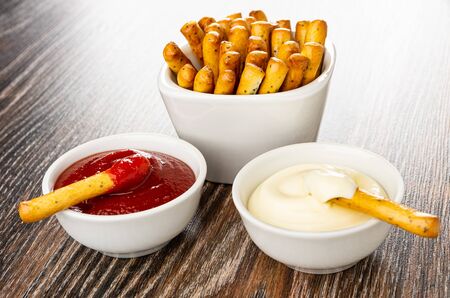 Bread sticks in white bowl, breadstick in bowls with ketchup and mayonnaise on dark wooden table