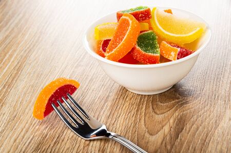Close up of marmalade on fork, multicolor marmalade in form slices of lemon in white bowl on wooden...
