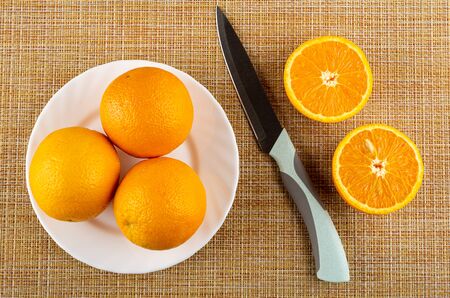 Full fresh oranges in white plate, kitchen knife, two halves of orange on brown mat. Top viewの写真素材