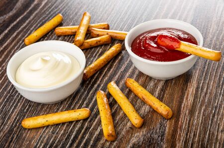 Scattered bread sticks, bowl with mayonnaise, breadstick in bowl with ketchup on dark wooden tableの写真素材