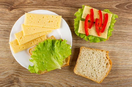 Slices of cheese, sandwich with lettuce in white plate, sandwich with cheese, leaves of lettuce, sweet pepper, slice of wheat bread on wooden table. Top viewの写真素材