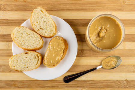 Slices of wheat bread, sandwich with peanut paste in white plate, teaspoon, jar with peanut butter...