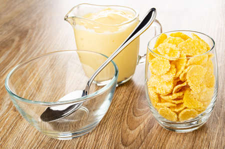 Spoon in empty bowl, pitcher with fruit yogurt, transparent glass with corn flakes on brown wooden...