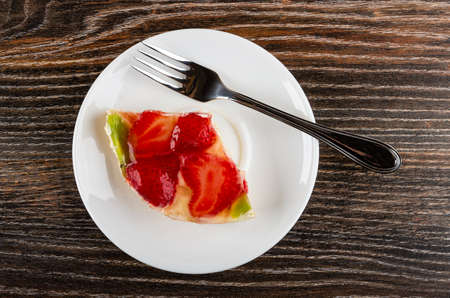Piece of pie with slices of strawberry and kiwi, fork in white plate on dark wooden table. Top view