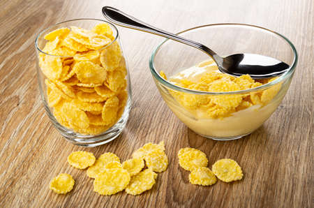 Glass with cornflakes, spoon in transparent bowl with corn flakes and fruit yogurt on brown wooden...