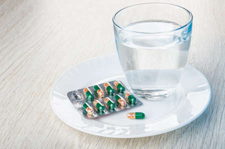 Blister of medicine in transparent capsules, single capsule, glass of water on white plate on wooden...