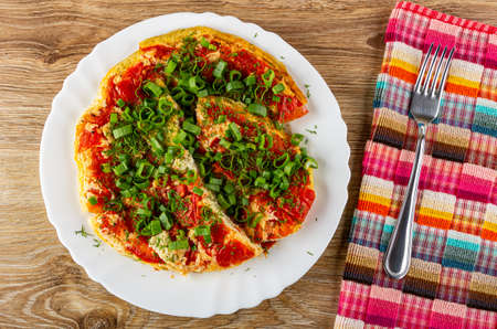 Pieces of cooked omelette with tomatoes, green onion, dill in white dish, fork on checkered napkin on wooden table. Top viewの写真素材