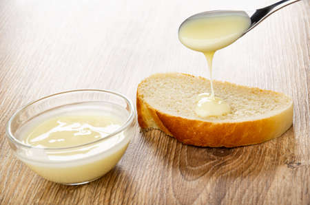 Transparent bowl with milk, poured condensed milk from teaspoon on slice of bread on wooden table