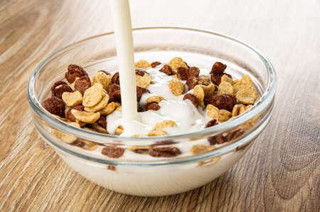 Pouring yogurt in bowl with cereal grains breakfast with chocolate and caramel on wooden table