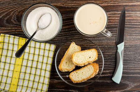 Teaspoon in bowl with sugar, checkered napkin, cup with fermented baked milk, pieces of sprinkled horn-shaped buns in transparent glass saucer on dark wooden table. Top viewの写真素材