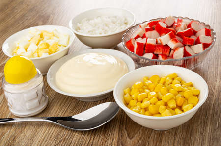 Prepared ingredients for crab sticks salad, spoon, salt shaker on wooden table