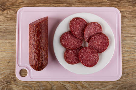 Piece of smoked sausage, slices of sausage in white plate on pink cutting board on wooden table. Top...