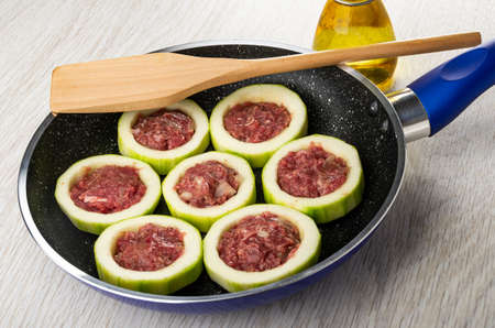 Rings of squash with filling from forcemeat, spatula in frying pan, bottle of vegetable oil on wooden tableの写真素材